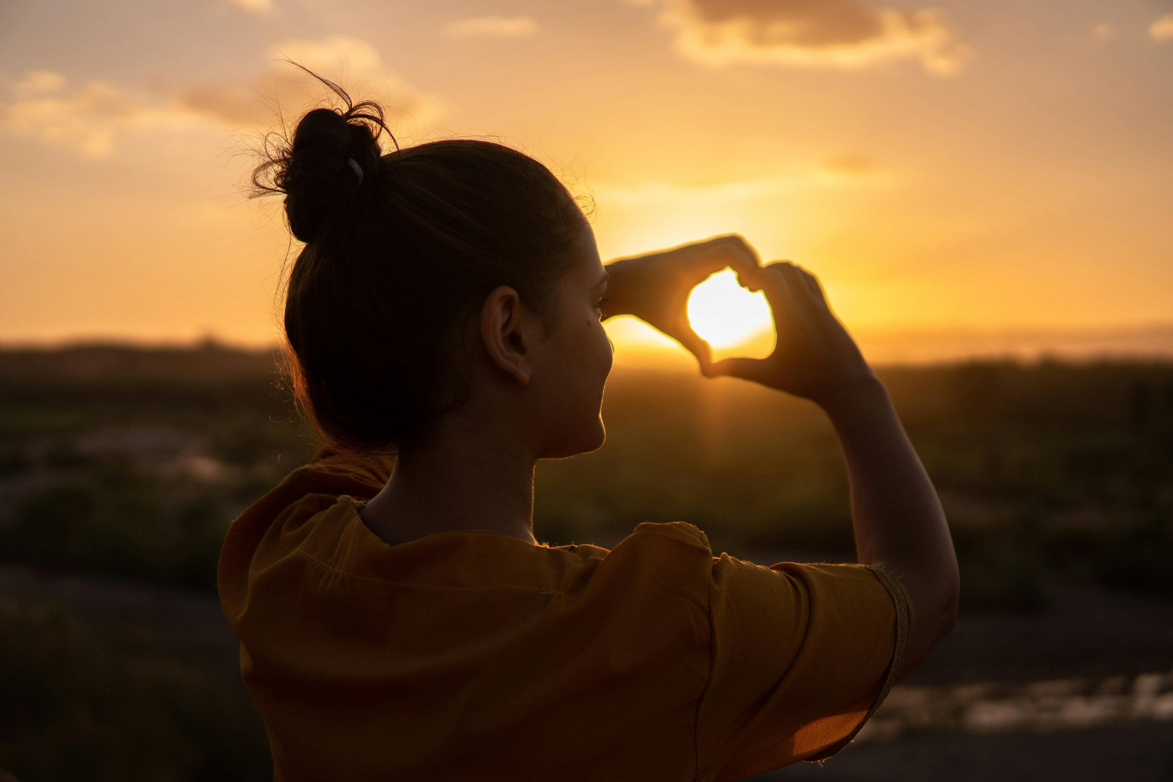 A woman stands facing a golden sunset with her back to the viewer, forming a heart with her fingers around the sun. The warm light symbolizes inner peace, self-love, and personal growth beyond surface-level self-care rituals.