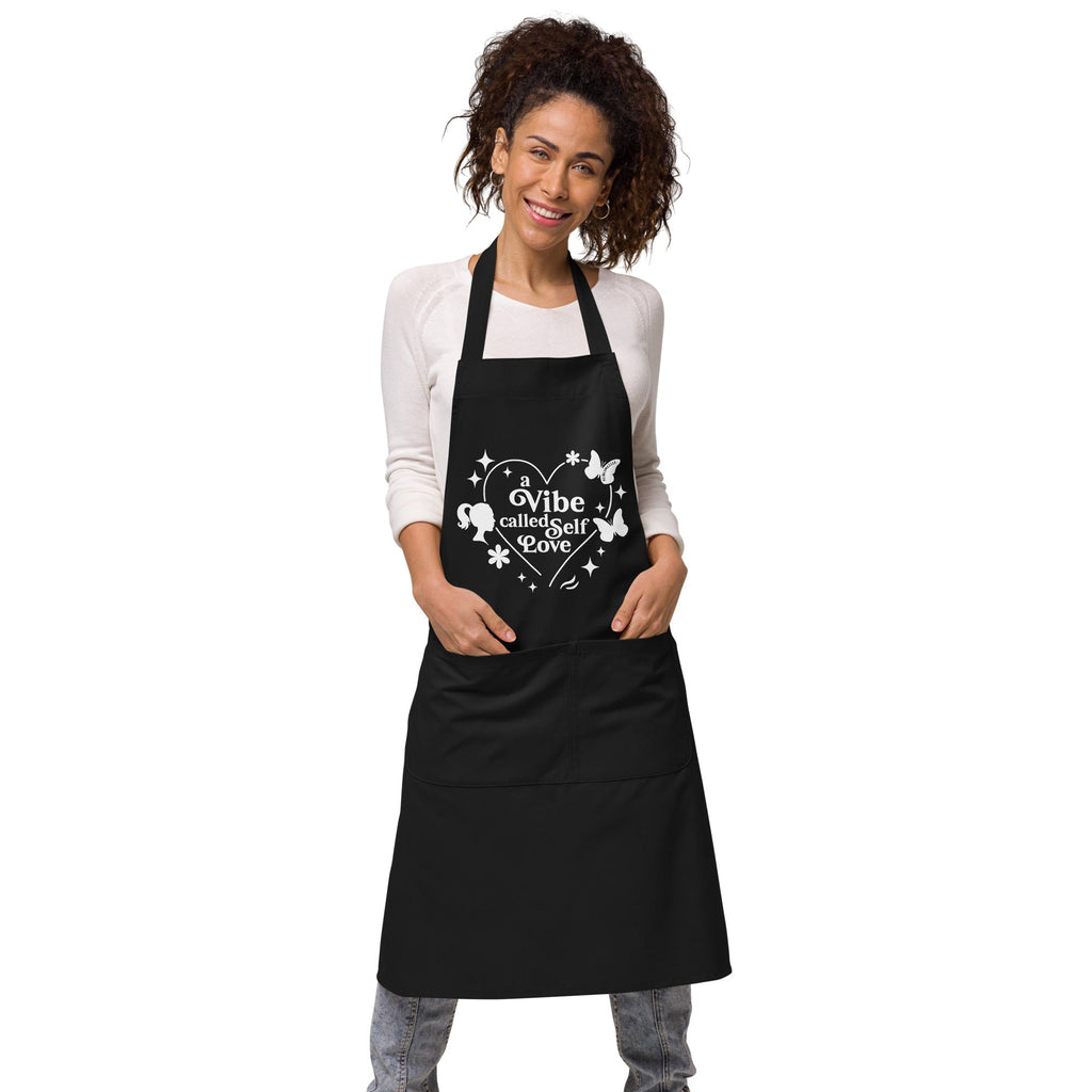  Woman wearing black apron with front pockets, showing ‘A Vibe Called Self-Love’ design, perfect for mindful cooking and self-care enthusiasts-SublimeElle Self - Love