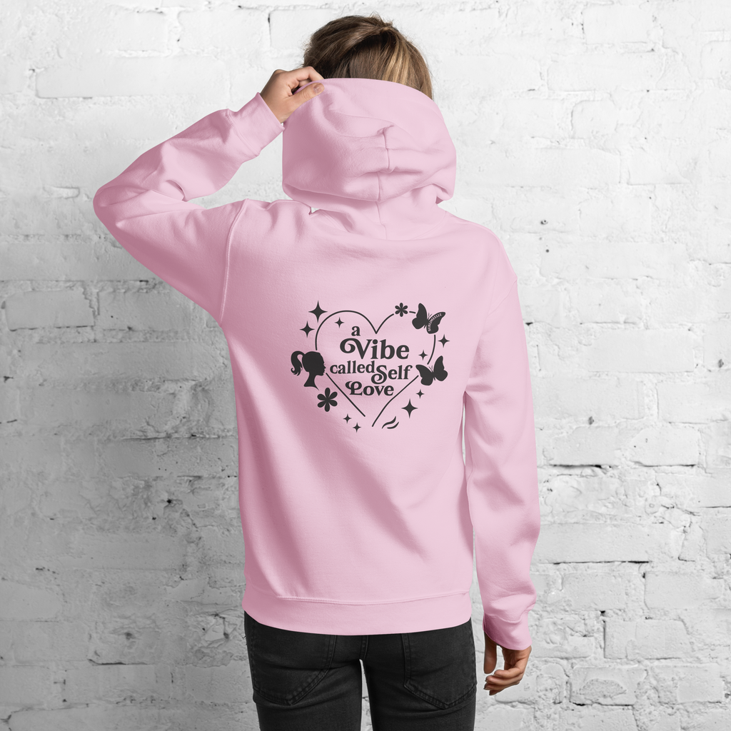 Woman standing facing a wall, wearing blush pink hoodie with quote 'a vibe called self-love' in the back. Self-love apparel 